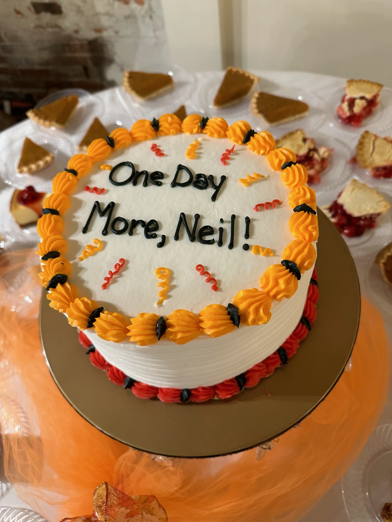 "A white cake with yellow and orange frosting details that reads "One Day More, Neil!""