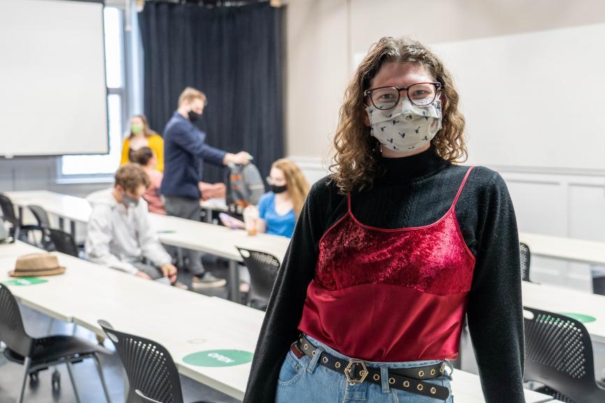 Student pictured in black long sleeves and glasses, wearing a mask, in front of a group of students working on a project