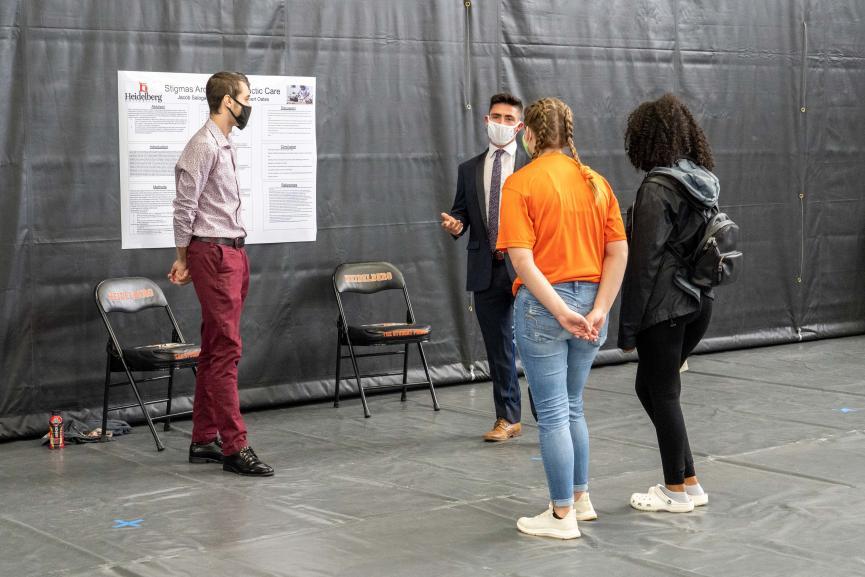 Two students present poster in open gymnasium to two students, back turned, all wearing masks and socially distancing