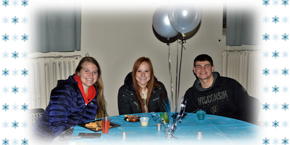 Three students sitting at a table with a snowflake tablecloth and balloons, eating hot pretzles and other snacks. On either side is a white border with blue snowflakes.