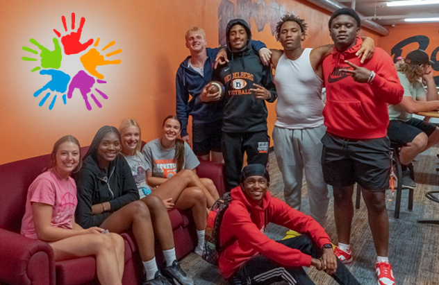 A group of several diverse students hanging out in the Brown basement lounge. The walls are orange and an image of 5 multicolor hands is superimposed.